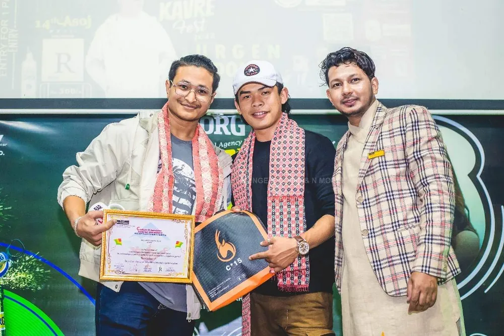 Three men on stage at a fashion event, one holding a certificate and the C-15 award plaque