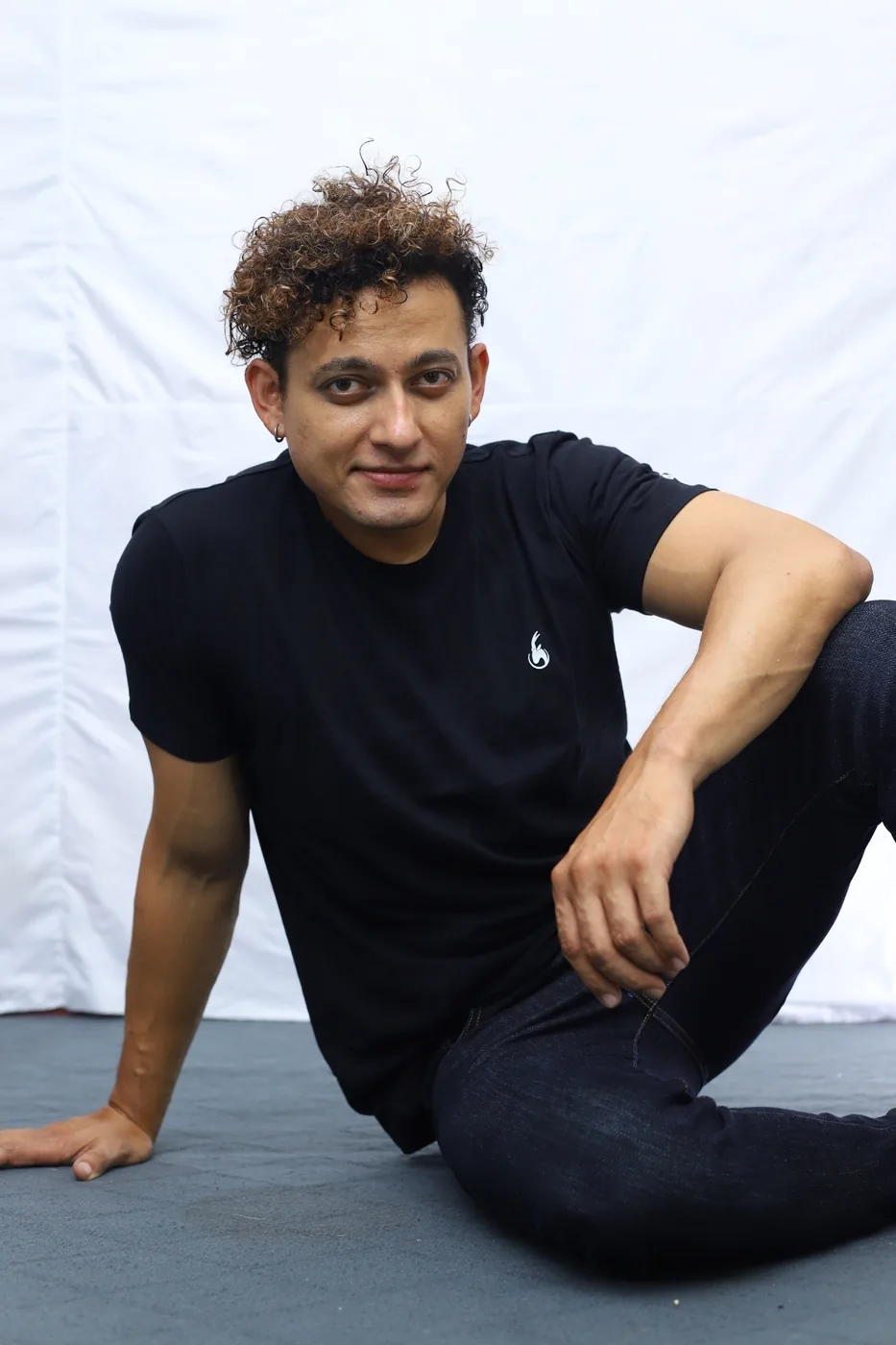 Model seated on the studio floor in a black C-15 t-shirt and dark jeans, smiling at the camera