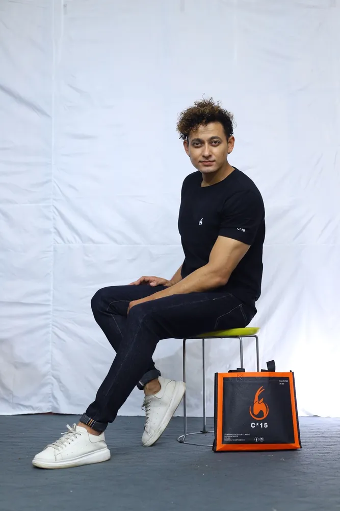 Model seated on a stool in a black C-15 tee and joggers with the orange C-15 tote bag beside him