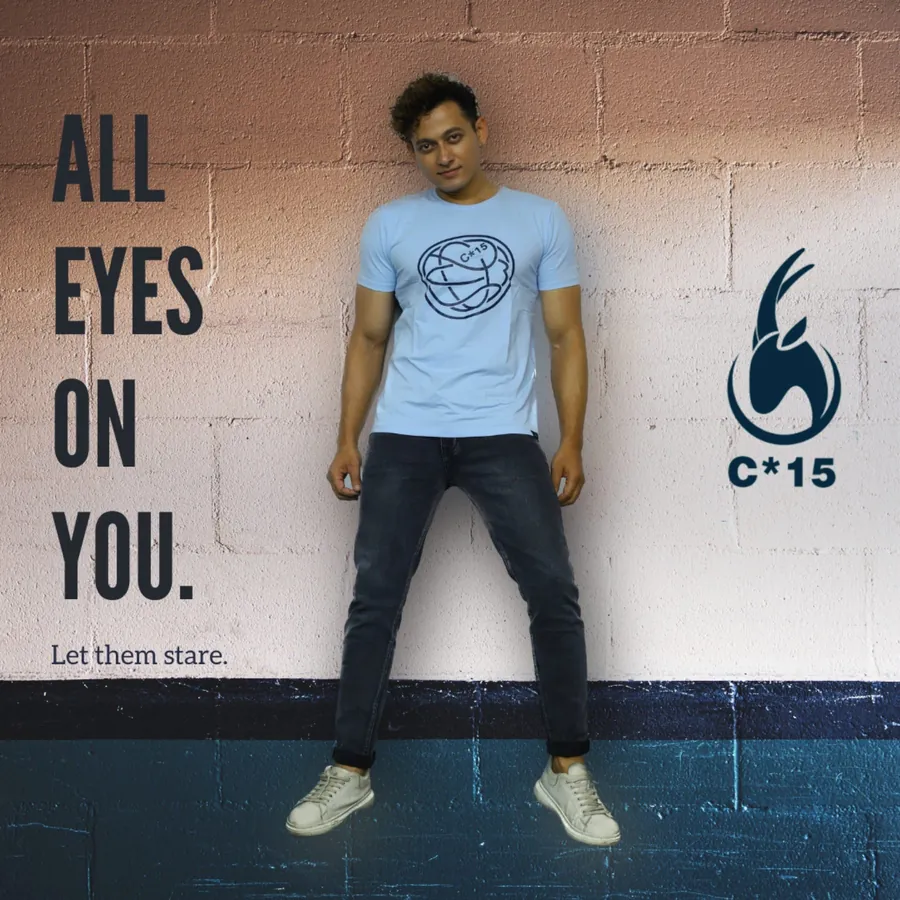C-15 campaign photo of a man in a light-blue graphic tee and dark jeans against a painted brick wall